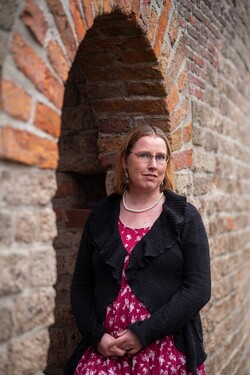 Foto van Lonneke, blanke vrouw met donkerblond haar, rode jurk met witte bloemen, zwart vest en parelketting in een nis van de Burcht in Leiden