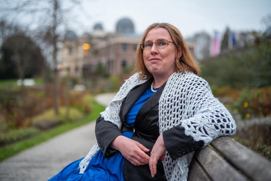 Foto van Lonneke: blanke vrouw op een bankje in een park met halflang blond haar, bril, koningsblauwe jurk, zwarte jas, lichtblauwe omslagdoek. Achtergrond is vaag, Sterrenwacht Leiden.